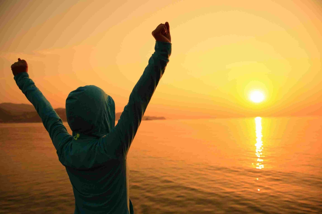 confident woman with arms raised gazing at the ocean at sunset